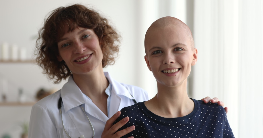 Smiling empathic 40s professional oncologist doctor in white uniform embracing shoulders of happy optimistic millennial female bald hairless patient, dreaming of full recovery, looking at camera. | Shutterstock HD Video #1063857421
