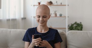 Happy sincere bald woman resting on sofa with smartphone in hands, communicating with cancer fighters, finding support in social networks groups, using healthcare applications, fulfilling life.