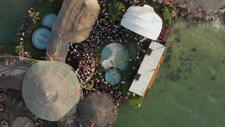 People dancing and partying at tropical Beach club Festival Party by the ocean in sunny weather with Palm trees and Pools, Aerial Birds Eye View Top Shot from above