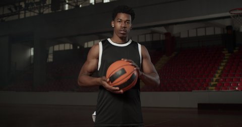 Video portrait of a basketball player bouncing a ball. African american man in uniform posing inside sports hall.