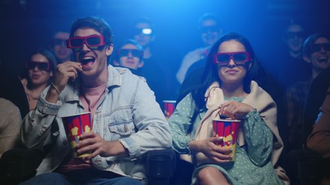 Cheerful friends having fun in cinema. Joyful couple laughing in movie theater. Happy woman and boyfriend eating popcorn in dark hall. Relaxed people spending weekend in cinema.