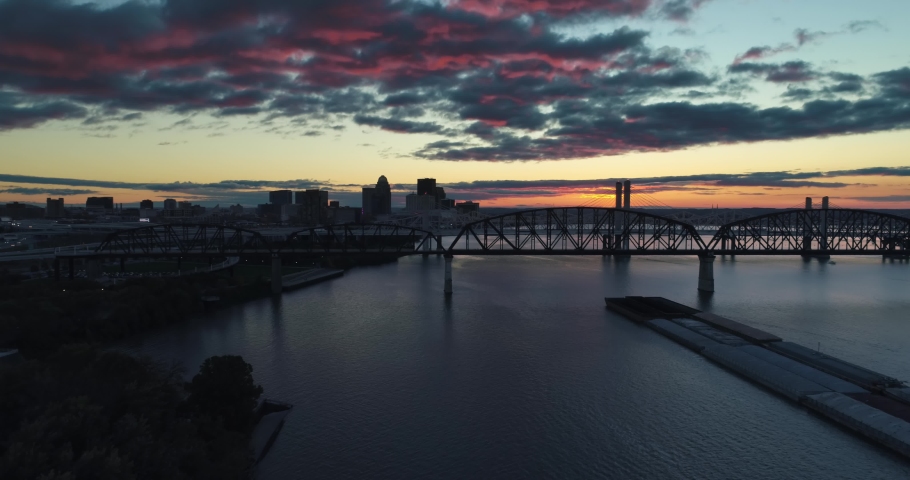 Downtown Louisville, Kentucky aerial overlooks city and Big Four Bridge at sunset. Colorful evening sky with cargo barge on Ohio River waterway.