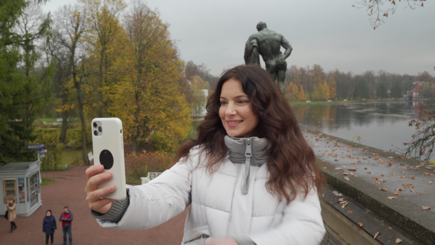 Happy pregnant woman in warm coat using smartphone camera in autumn park. Brunette woman with long hair in Catherine Park, Tsarskoye Selo, Saint Petersburg, Russia.