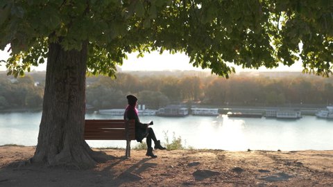 Solo activity and social distancing. Concept of time spending alone. Silhouette of an attractive woman, sitting alone in park bench, wearing fashion outfit, surfing in internet on her gadget.