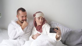 Couple on bed take photo on smartphone, adorable female hold mobile phone in hands, posing at camera, have rest.