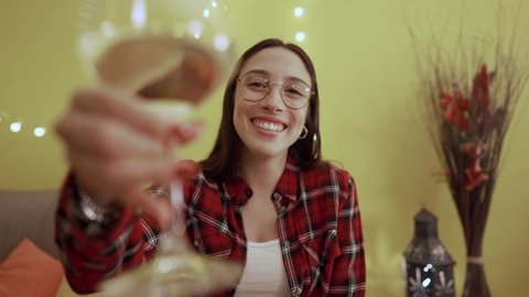 Young Caucasian women POV talking making a toast holding a champagne glass looking at camera. Single smiling female person using video conferencing technology to celebrate online for social distancing