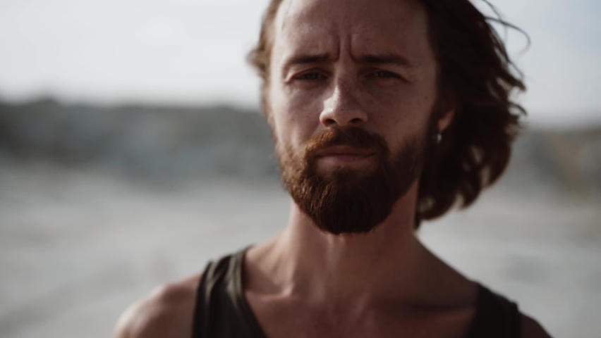 a man who looks like a tramp with long hair and a beard looking at the camera, turns his head, hair fluttering in the wind, against a blurred background, slow motion Royalty-Free Stock Footage #1063884106