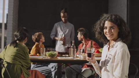 Medium shot of young mixed-race woman with dark curly hair is sitting at dining table with wine glass in her hands, looking at camera while her friends talking on background