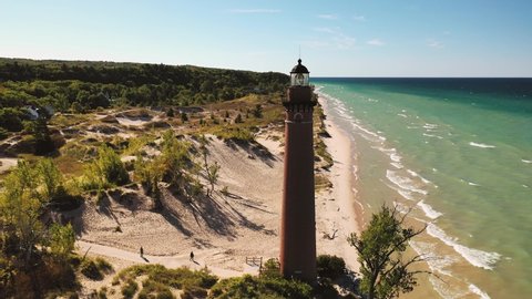 Beautiful aerial view circling around Little Sable Lighthouse along the shoreline of Lake Michigan sitting amongst vegetation and tree covered sand dunes with a clear blue sky above.