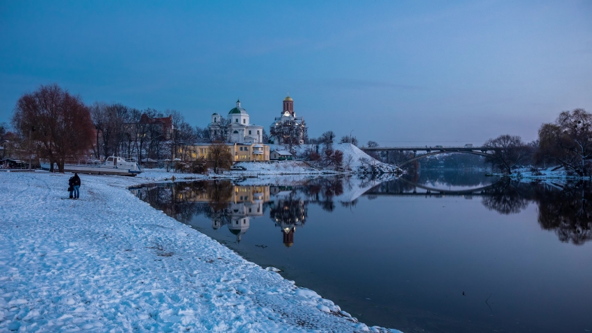 Beautiful winter time-lapse. A loving couple stands on the river bank. Snow. The water flows. Churches and bridge in the evening in the city.