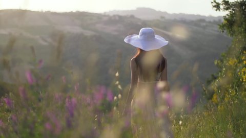 Beautiful young woman with summertime dress walking through meadow in summer. Romantic sunset behind hills during sunny day in spring creating relaxing, warm and natural atmosphere. Central Italy, 4K.