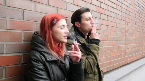 Bad habit young people couple stand smoking cigarettes exhaling dense smoke communicating near the wall