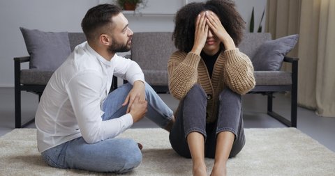 Upset young family couple african american girl black wife sitting on floor holding her head with hands worried about resentment caucasian man screaming waving her hands in anger, quarrel concept