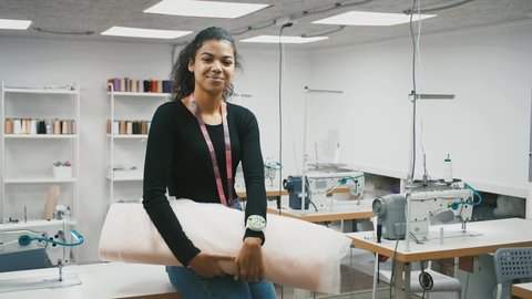 Young mixed race lady, tailor with tape measure on neck is holding roll of tulle fabric and smiling, posing sitting on workstation equipped by sewing machine at modern sewing workshop. Slow motion