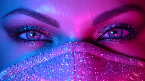 Womans face in glitter protective mask, amazing different make-up under neon colorful light. Female with shining shadows. Glamour, art, beauty, fashion, pandemic, covid-19 concept.