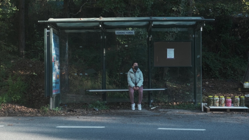 Vehicles Passing By A Girl In Face Mask Sitting And Waiting On Bus Stop During COVID-19. - wide shot