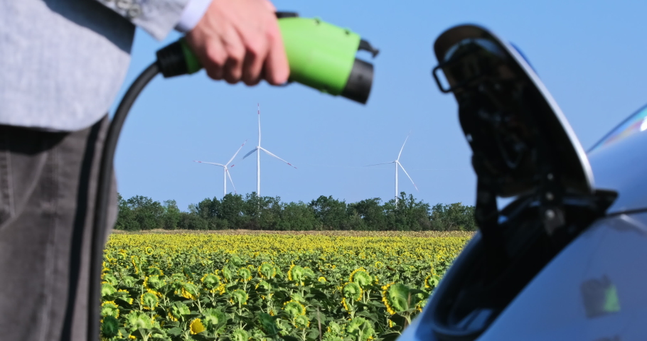 A man connects an electric car to a charger and several wind generators stop in the background. Overload in the electrical network due to electric cars. Royalty-Free Stock Footage #1063916383