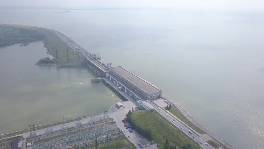 Novosibirsk Hydroelectric Power Plant is a hydroelectric power station on the Ob River in the Soviet district of the city of Novosibirsk. The aerial footage captures the full length of the concrete dam structure stretching across the wide reservoir, with the power station building positioned at the center. A road runs along the top of the dam, with vehicles visible traveling in both directions. To one side of the structure lies an extensive electrical substation with rows of transformers and transmission towers, while the other side opens onto the vast expanse of the Novosibirsk Reservoir. The surrounding landscape is flat and green, with forested banks curving along the shoreline. The water surface is calm and muted under overcast summer skies, giving the scene a functional, industrial atmosphere. The camera maintains a steady high-angle perspective, slowly tracking along the dam to reveal its scale and integration into the Siberian landscape. Construction of the station began in 1950, and it remains the only hydroelectric power station on the Ob River.