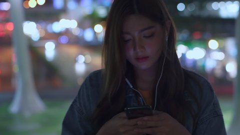 Young beautiful Asian woman walking in city street at night with listening to broadcast online music on headphones from smartphone. Pretty girl relax and enjoy with radio streaming from mobile phone