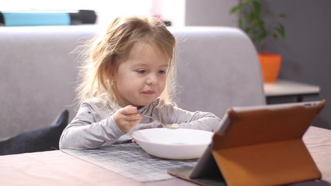 pretty little blonde girl in pajama eating milk porridge with asterisk vermicelli, looking in the tablet. breakfast, morning home. FullHD footage.