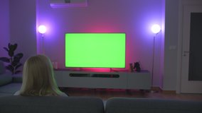 Lonely girl sitting on sofa in dim purple and pink lights in living room watching TV big green screen
