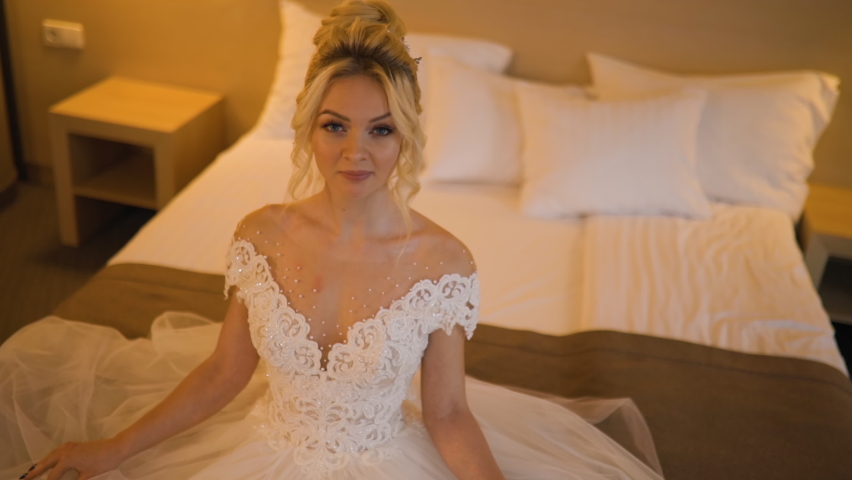 Cute blonde bride in wedding dress sits in hotel room and looks at you