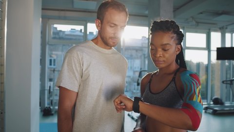 African american woman athletic and her coach trainer sport doctor standing in the gym center and looking at the sport results at fitness tracker smartwatch while prouding of personnel record