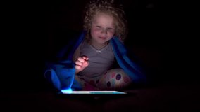 A charming curly-haired girl of four years old sits in bed under the blanket before going to bed and makes a video call with her parents and laughs out loud.	