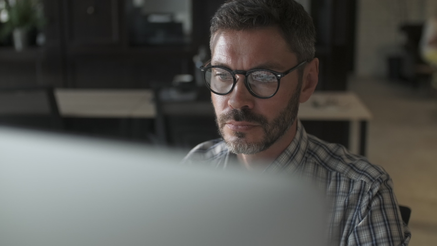 Close Up Young Man in Fashion Glasses Eye Looking Monitor, Surfing Internet. Stylish Male Working With Laptop or Tablet From Home in Him Home Office. The Monitor Screen Is Reflected In The Glases