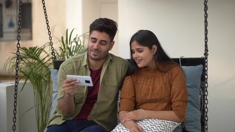 Two Indian friends, couple watching funny videos using smart phone laughing together. Cafe Exterior. Indian couple watching funny social media content while enjoying on swing.