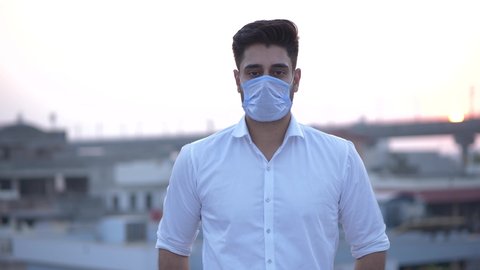 Close-up shot of a young Indian adult male looking at camera standing on a roof with metro city background. Close-up shot of an Indian man looking at camera wearing face mask while standing outdoors.