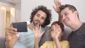 Positive homosexual parents and kid using cell for video call, waving hello, making like gesture and smiling, pointing at screen. Happy family and communication concept