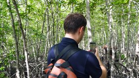 Young Smiling Man Tourist with Backpack Makes Video on Smartphone in Mangrove Forest. Traveler Shooting Stories Clip Movie on Phone for Social Media on Wooden Walkway in Sunny Woods.