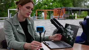 Girl blogger blogging in a cafe on a summer terrace outdoors. A blogger mom with a baby in a pram shoots her vlogs and short videos on an action camera on a tripod and stylish laptop on a table