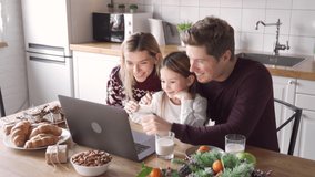 Happy family young parents with cute small kid child daughter holding xmas gift waving hands using laptop computer at home sit at Christmas table having virtual party dinner on video call together.