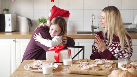 Happy dad wearing santa hat giving excited cute small kid daughter xmas gift sitting at kitchen table. Family parents preparing present for child girl hugging celebrating Christmas holiday at home.