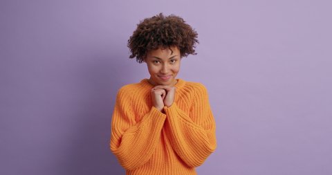 Positive dark skinned young woman with Afro hair looks pleasantly at camera keeps hands under chin smiles gently feels delighted and shy poses against purple studio wall. Happy face expressions