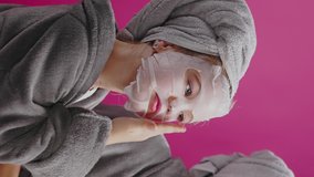 Vertical video portrait young teenager girl pretty child wearing sheet mask with her mother looking into mirror at pink background. Mom and daughter. Cosmetics.