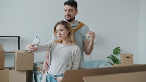 Young man and woman are taking selfie with keys to new apartment after relocation. Young people are kissing and hugging using modern smartphone camera.