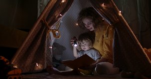 Beautiful caucasian woman taking care of her child. Young mom is reading book of fairy tales for kid. Family in cozy tent in bedroom in evening 4k footage
