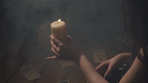 Woman performing mystical esoteric ritual in dark room with smoke. Fortune-teller doing purification act with candle in hand and tarot cards on table. Female predicting destiny and forecasting future.
