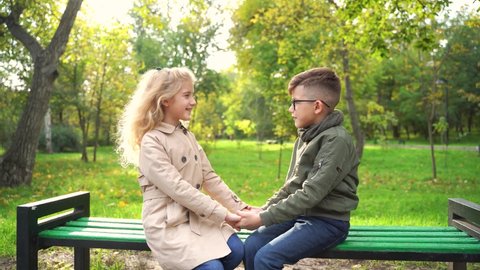 Little couple sitting on bench in autumn park. Happy boy and girl smiling and looking at each other while sitting on bench during break in autumn park