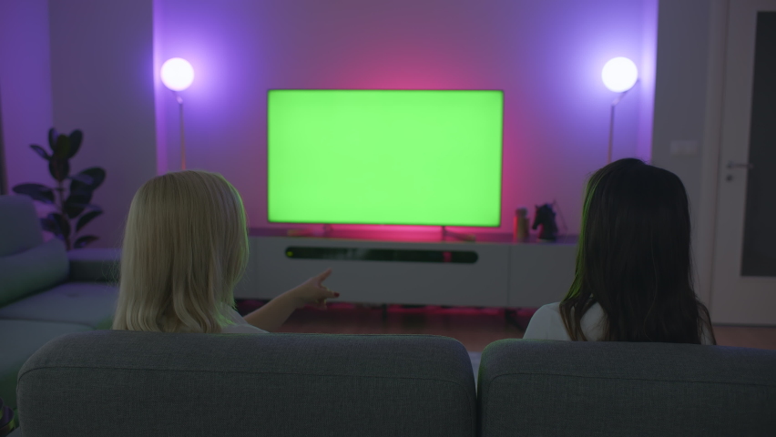 Dim living room lit by colored lights one blonde one brown hair young women talking about movie or tv series big screen television in the background | Shutterstock HD Video #1063964839