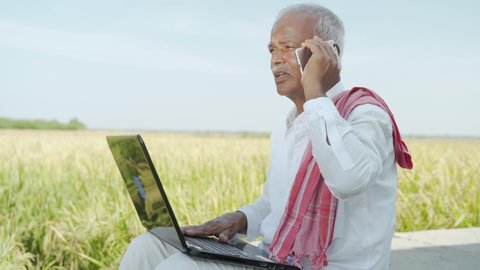 Indian farmer talking on mobile phone while busy looking into laptop near the agriculture farmland - concept of farmer using technology, internet in rural India.