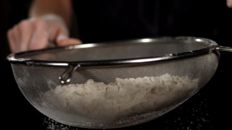 Close-up particles of white flour is falling through a steel sieve on black background in slow motion. Baker is sifting flour. High quality FullHD footage