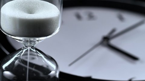 A white hourglass made of glass, in the foreground with an out-of-focus analog wall clock in the background.