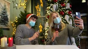 Couples sit at the table with Corona masks on Christmas with a smartphone and clash with wine glasses on a video call.