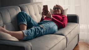 A cheerful young woman in glasses taking a video call with her smartphone while lying on a couch in the living room.