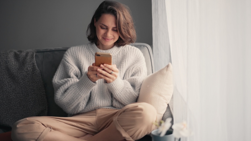 Relaxed young woman using a smartphone and drinking coffee from a mug while sitting on the sofa. Millennial girl spending time at home with cell gadget technology. Royalty-Free Stock Footage #1063974649