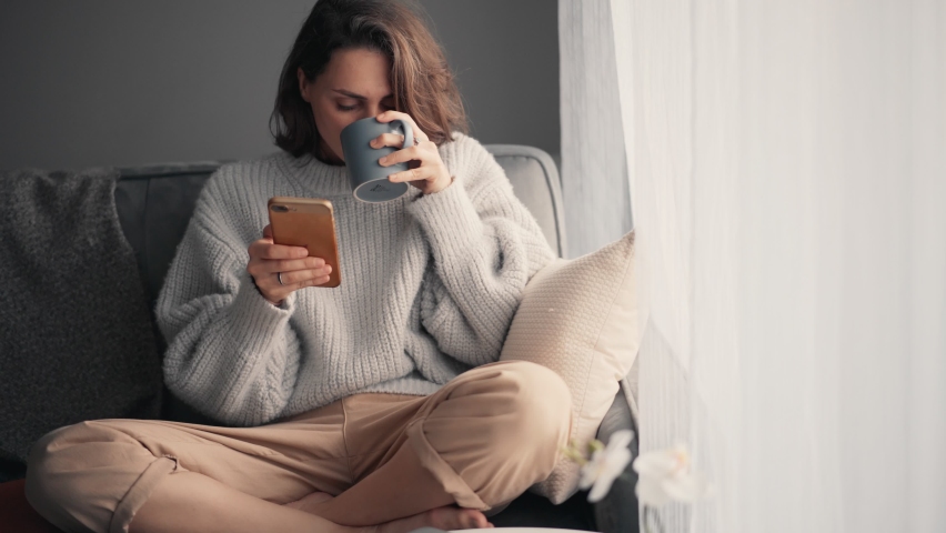 Relaxed young woman using a smartphone and drinking coffee from a mug while sitting on the sofa. Millennial girl spending time at home with cell gadget technology.